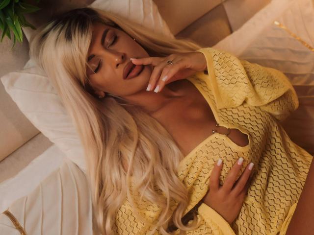SalmaRoze with long blonde hair, eyes closed, wearing a yellow crochet top, touching her lips, hand on her chest, nails painted white, lying on a bed.