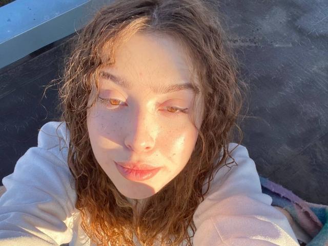 El rostro de Jessika Bernskoetter, con cabello castaño rizado, pecas y piel clara, lleva una camisa blanca, delineador de ojos y está mirando hacia abajo en un selfie tomado en un día soleado y con luz cálida..