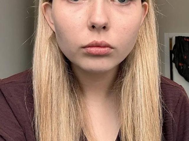 JoneGann has pale skin, straight blonde hair, and a serious expression with pinkish lips. She's wearing a dark maroon shirt, and her nose is slightly upturned.