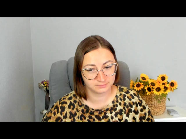 PaesAnda with glasses, brown bob, and leopard print shirt, sitting in a gray chair, smiling slightly, with a sunflower basket on a white table to the right.