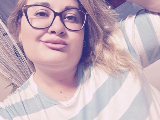 RadaRori with glasses, smiling, wearing a blue and white striped shirt, has a small mole on her chin, blonde hair, and hoop earrings, in a selfie.