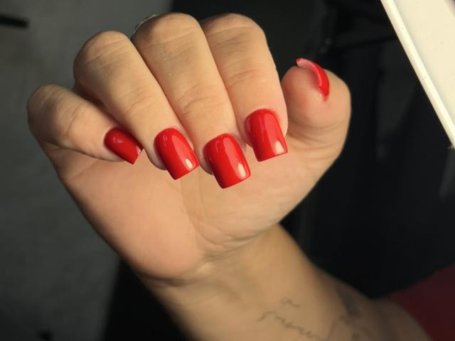 RadaRori's hand with shiny red nail polish, showing off her well-manicured, glossy, medium-length nails. Tattoo visible on her wrist.