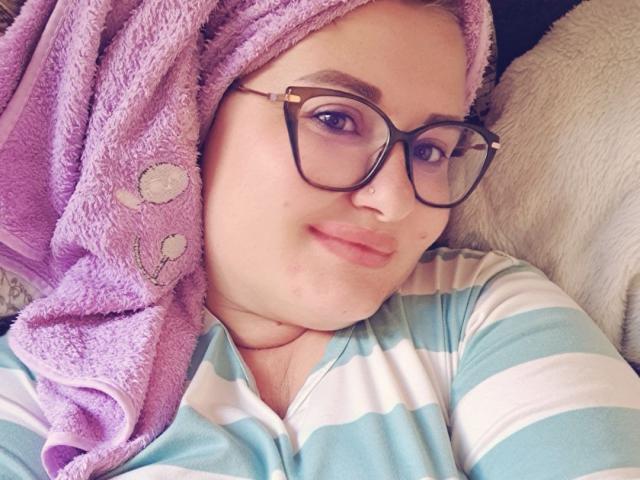 RadaRori has a purple towel on her head, wearing glasses and a striped shirt, with a slight smile and a pimple on her cheek. She's looking at the camera.