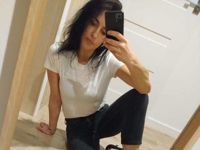 CelesteSommer, con i capelli neri, una maglietta bianca e pantaloni neri, seduta sul pavimento di legno, che si scatta un selfie allo specchio con un telefono nero, con il braccio destro appoggiato, il ginocchio sinistro sollevato e il ginocchio destro a terra..