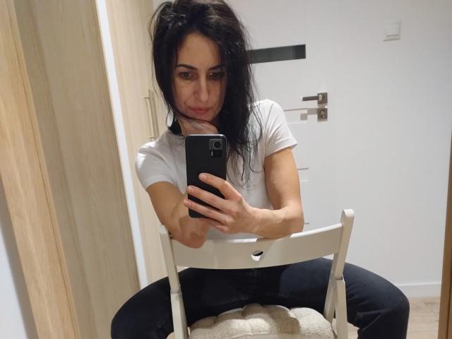 CelesteSommer mit dunklem, zerzaustem Haar, weißem T-Shirt, schwarzen Hosen, sitzend auf einem weißen Stuhl, macht ein Spiegel-Selfie, hält ein schwarzes Telefon, lächelt leicht, sichtiger Hintern, kleines Gesäß, Holztür im Hintergrund..