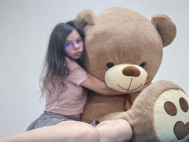 VikkiRi with long, wavy black hair, glasses, pink shirt, and checkered skirt, hugging a huge brown teddy bear, sitting against a plain white wall.