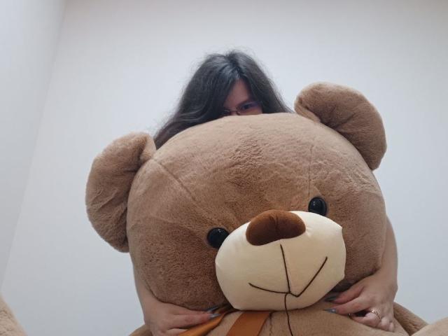 VikkiRi with long dark hair, partially hidden behind a huge brown teddy bear, gripping its furry face. Her blue-painted nails and ring are visible.