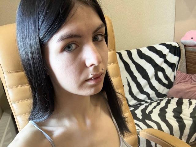 NobukoGraue, con la pelle chiara e i capelli neri e lisci, ha un piercing al naso, occhi azzurri e indossa una canotta grigia, seduta su una poltrona in pelle color caramello..