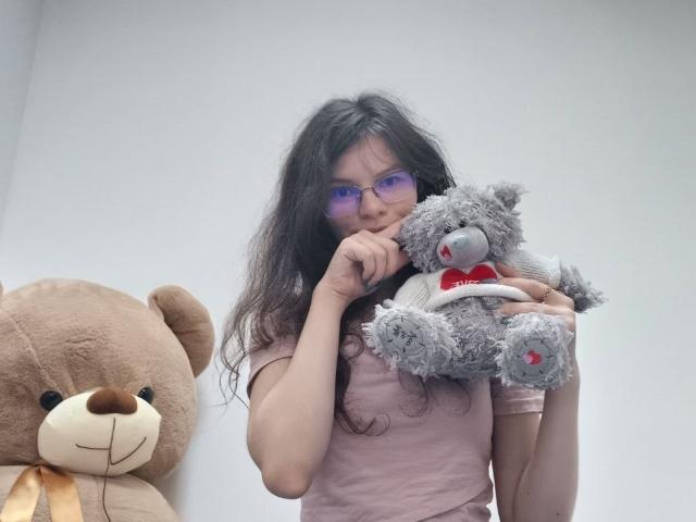 VikkiRi with curly black hair, glasses, and pink shirt, holding a fluffy grey teddy with a red bow, standing next to a large brown teddy.