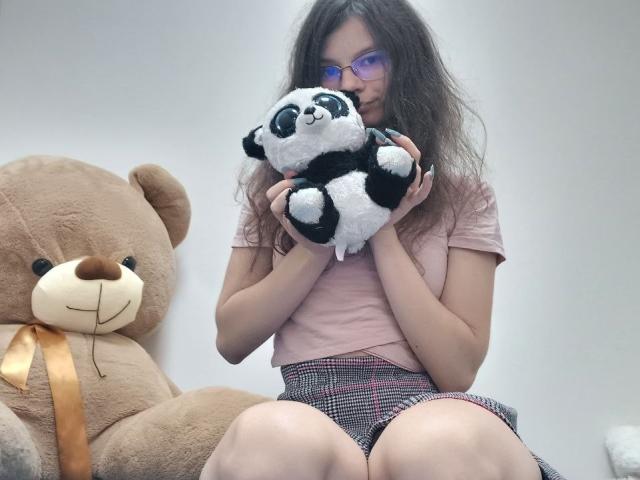 VikkiRi with long brown hair, glasses, pink top, and plaid skirt, holding a black-and-white stuffed panda, sitting in front of a large brown teddy bear.