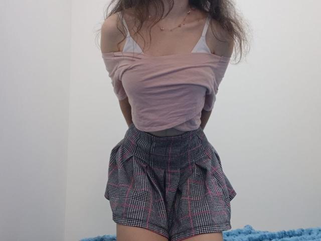VikkiRi with off-shoulder pink top, white bra, and plaid shorts, arms behind back, showing tits, small waist, and fluffy blue blanket bottom right.