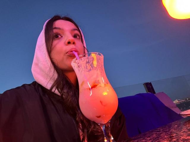 Molray, avec de longs cheveux noirs, un sweat à capuche blanc, sirotant une boisson rose et glacée avec une paille, un ciel crépusculaire bleu en arrière-plan, une lumière orange en haut à droite..