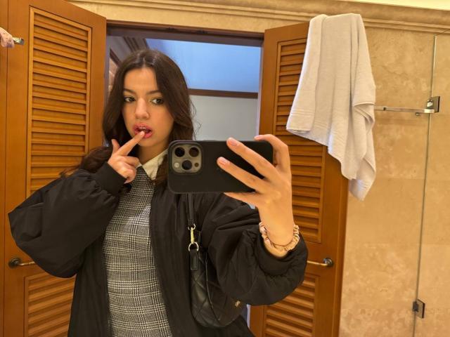 Molray, com cabelo castanho comprido, jaqueta preta e camisa xadrez cinza, chupando o dedo, tirando uma selfie com um celular preto, uma toalha branca pendurada ao fundo..