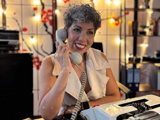 Curly-haired JuliaNewell with gray hair, smiling, holding a silver phone, wearing a white sleeveless shirt, sitting at a desk with a typewriter, looking happy and relaxed.