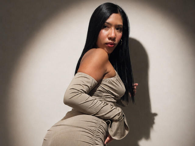 GabiHott con lunghi capelli neri, vestito scuro a spalle scoperte, che guarda oltre la spalla, ombra sul muro, mano sul fianco, che mostra glutei e seno, espressione seria, illuminazione soffusa..