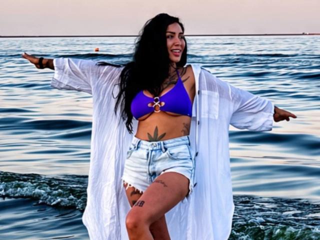 NyraNoble stands in the water, arms out, wearing a purple bra, white open shirt, and denim shorts. She has long black hair, tattoos, and a smile.