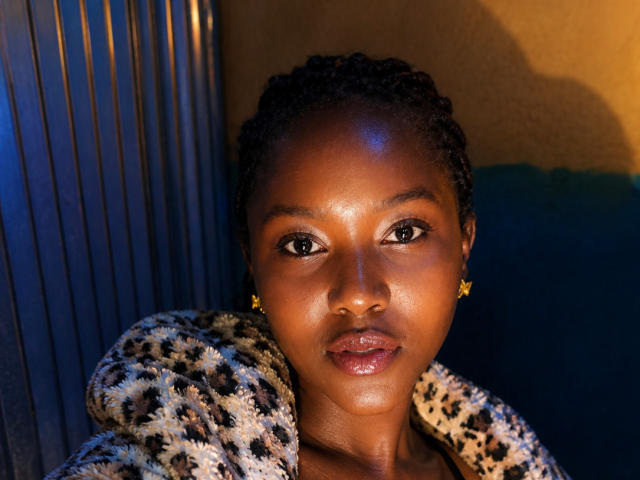 KapsLovie with dark skin, shiny lips, and star earrings, wearing a leopard print hoodie, has braided hair and a serious expression, illuminated by warm and blue light.