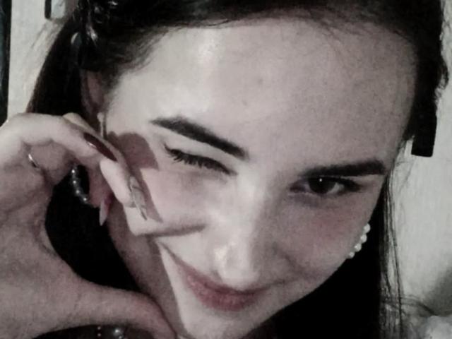 LupeEchavarria's face, winking with dark eyebrows, has pale skin and long dark hair, wearing pearl earrings, smiling subtly, hand touching her ear with a ring on her finger.