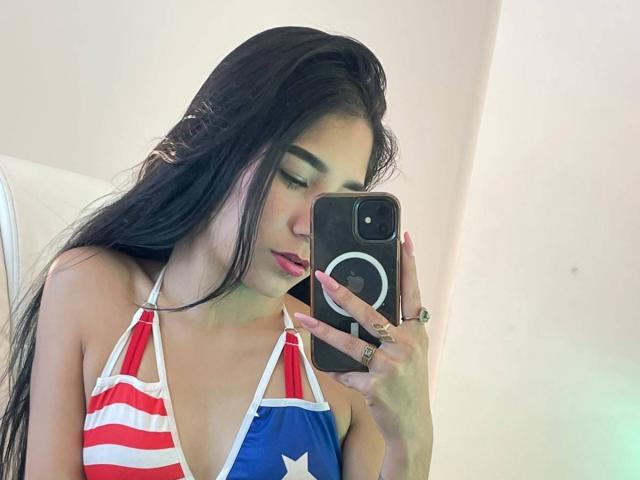 VioletaVelvet with long black hair, wearing a USA flag-themed halter top, taking a selfie with a black phone, pink nails, and red lipstick, in a mirror.