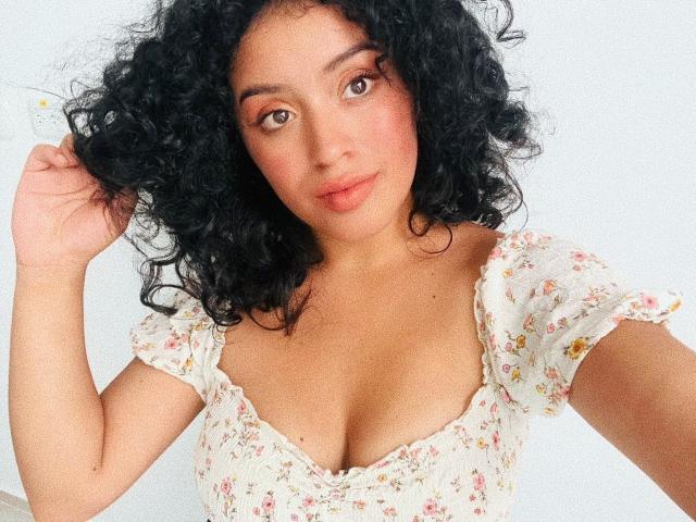 AlinnaPalacios with curly black hair, brown eyes, and pinkish blush, wearing a floral, low-cut, short-sleeve top, showing her tits, holding her hair with a gentle smile.