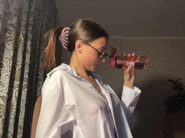 KassandraDalal with glasses, brown ponytail, pink scrunchie, white shirt, nipples visible, drinking from a pink water bottle, standing in a dimly lit room.