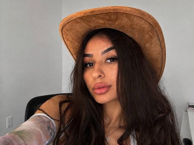 Emma Noor, met lang donker haar, bruine ogen en volle lippen, draagt een beige hoed, een kruisbeeldketting en een transparant shirt dat haar borsten laat zien. Haar wenkbrauwen zijn dik..