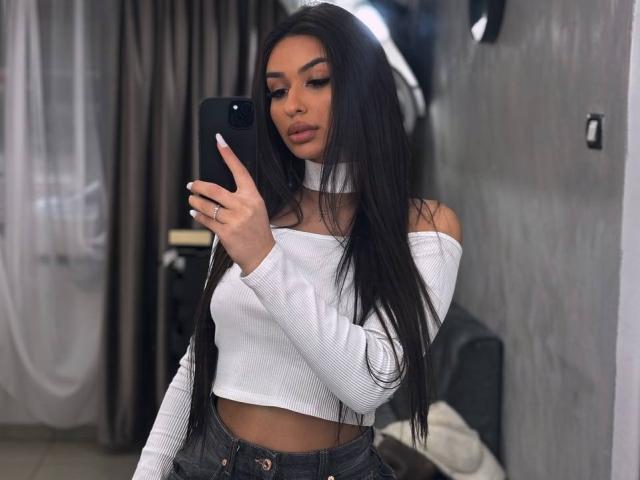 Emma Noor, com cabelos longos e pretos, blusa branca com um ombro à mostra e calça jeans, tirando uma selfie no espelho, mostrando seus pequenos seios, em um quarto com pouca luz..