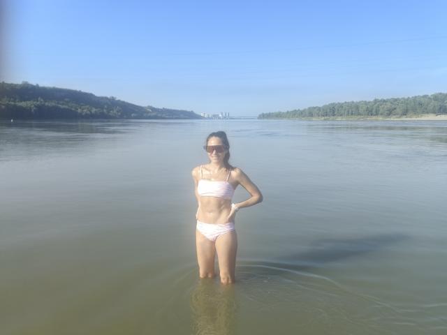 Miss Kinney staat in een rivier, gekleed in een roze bikini, met haar handen op haar heupen, en laat haar slanke, getoonde lichaam en kleine borsten zien. Ze draagt een zonnebril, op een zonnige dag..