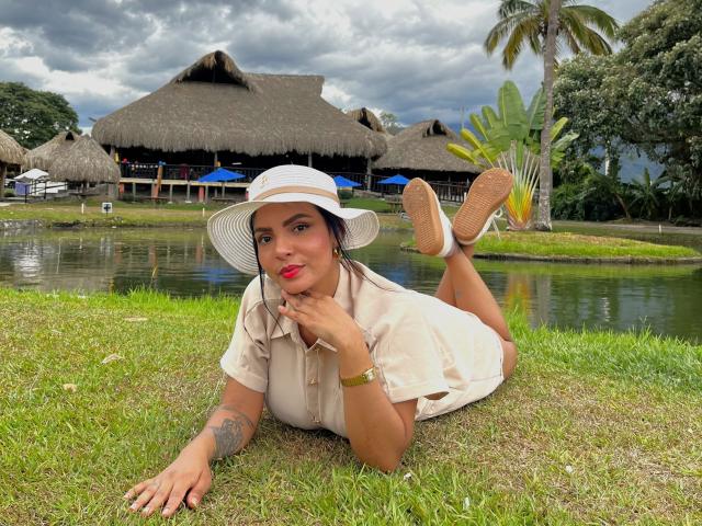 JessicaFiore lying on grass, wearing a white hat, beige shirt, and shorts, with her ass up and toes in the air, red lipstick, tattoo on left arm, yellow shoes.