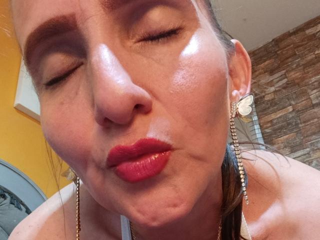 Het stralende gezicht van RubiAnderson, met gesloten ogen en rode lippen die alsof ze een kus uitdelen, draagt vlinderoorringen, en je ziet haar rimpels en een lichte glans op haar huid..