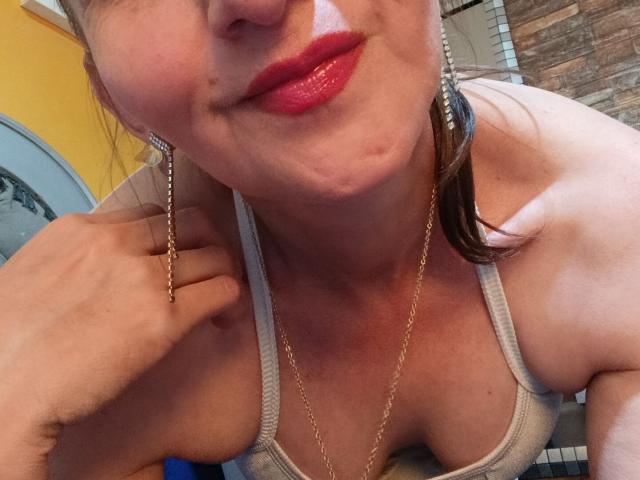 Het gezicht van RubiAnderson, met rode lippenstift, licht gezwollen wangen, een grijze tanktop, een gouden ketting en oorbellen, terwijl ze haar oor vasthoudt met haar hand. Borsten zichtbaar..