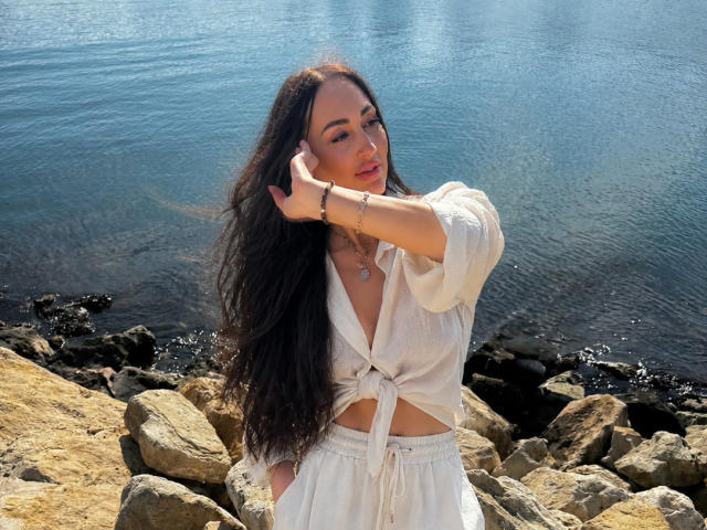 FreyaHott69 con cabello largo y oscuro, blusa blanca anudada y falda blanca, tocándose la cara en una orilla rocosa, agua azul en el fondo, luz solar, ligeramente sonrojada, pulsera en la muñeca..