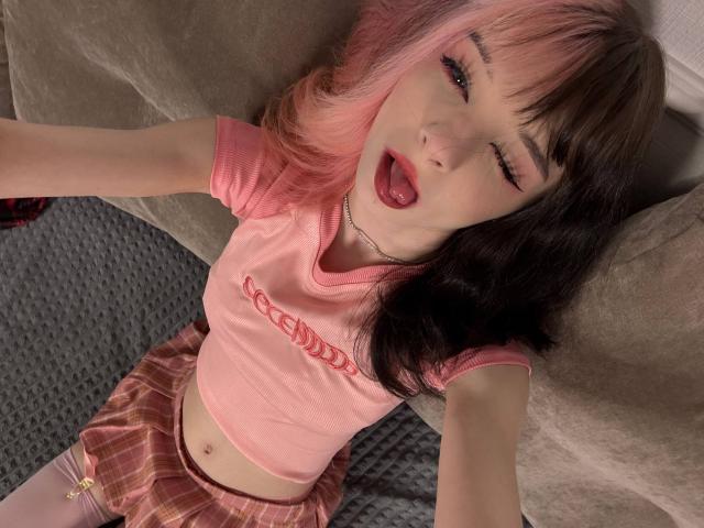 ChangCaddle with pink and brown hair, winking, tongue out, in a pink top and pink plaid skirt, lying on a carpet, wearing a silver necklace.