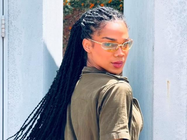 NaomiSteward with long black dreadlocks, wearing a green jacket and yellow glasses, looking over her shoulder with a confident smile, sunny outdoor setting.