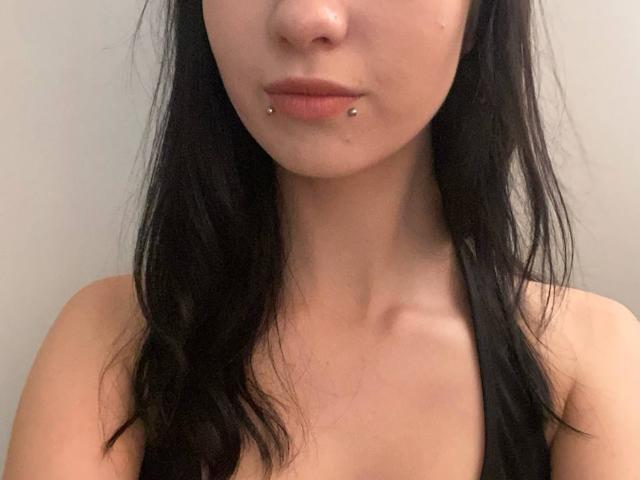 MelvaBhatt has black hair, pale skin, and a pierced lower lip with two small silver studs. She's topless, showing her bare shoulders and upper chest.