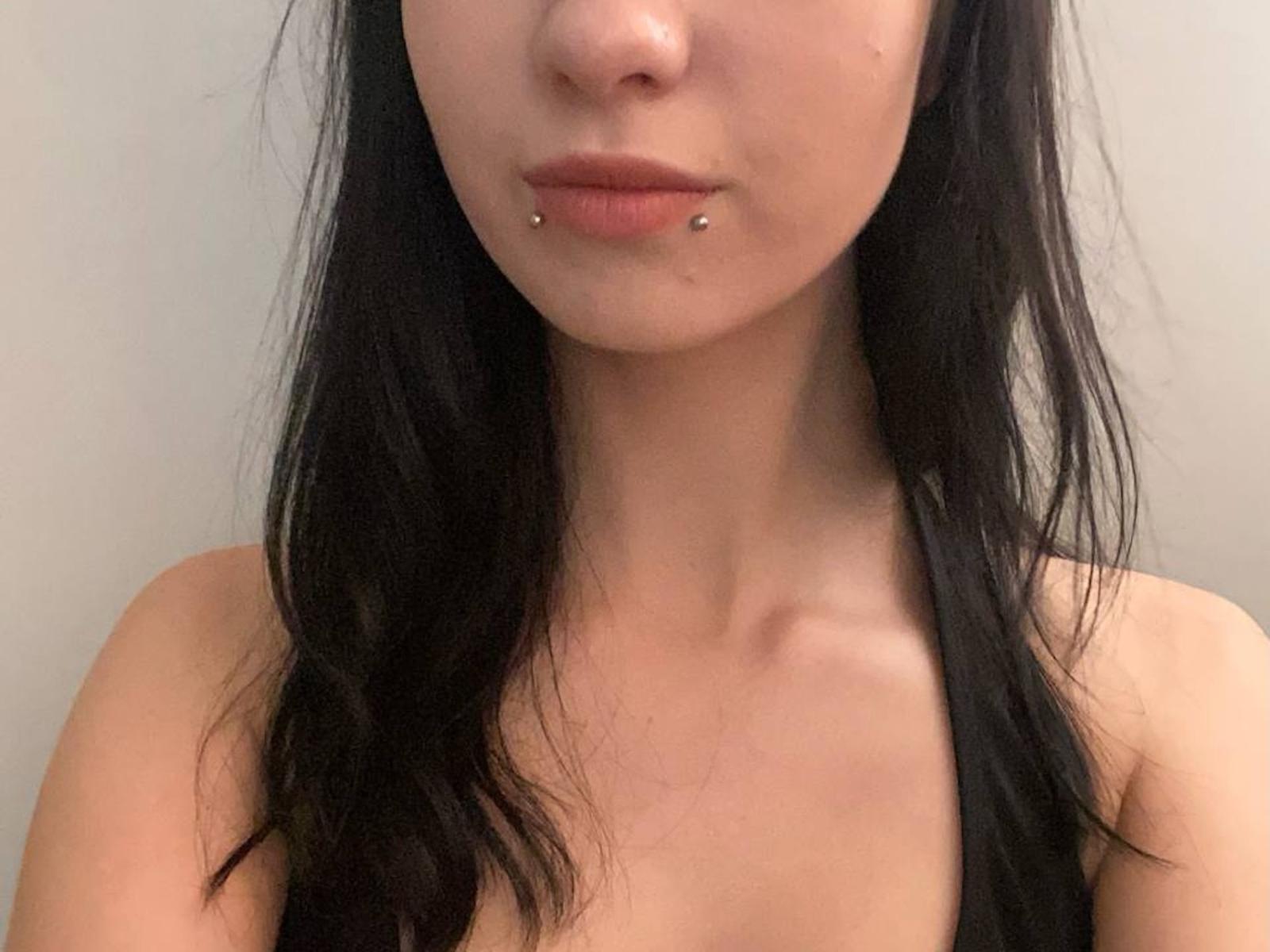 MelvaBhatt has black hair, pale skin, and a pierced lower lip with two small silver studs. She's topless, showing her bare shoulders and upper chest.