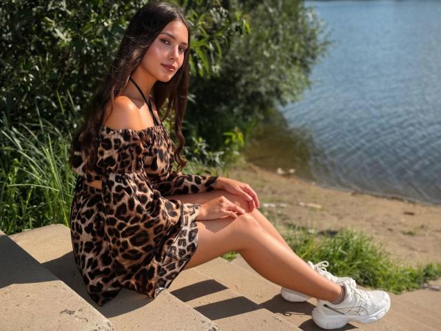 Stephanies, mit langen, dunklen Haaren, trägt ein Kleid mit Leopardenmuster und sitzt auf einer Steinkante, die Beine gekreuzt, weiße Turnschuhe, blickt leicht zur Seite, in der Nähe eines Gewässers..