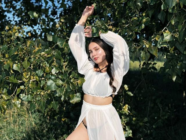 Stephanies-hot with long brown hair, wearing a white sheer crop top and skirt, arms raised, standing in front of leafy green branches, sunlight highlighting her tan skin and pink lips.