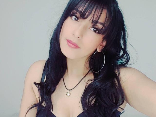 Hotcaroline33, con lunghi capelli neri, orecchini a cerchio, rossetto rosa e una collana a forma di cuore, mostra il suo seno con una maglietta nera. Sta guardando la fotocamera con un sorriso carino..