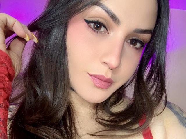 ElinaJhonson has pink lipstick, dark eyebrows, and eyeliner, with long brown hair and yellow nails, wearing a red lace top, showing a tattoo on her shoulder.