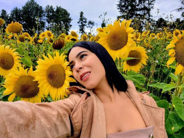 MarianaJackson with black hair, tan jacket, smiling, holding out arm, surrounded by sunflowers, showing her tits, in a sunflower field, trees in the background, sunny day.