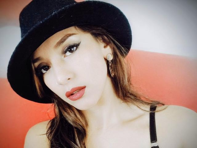 Hotcaroline33, con un cappello nero, capelli castani e rossetto rosso, indossa bretelle nere, ha un viso carino e piccoli seni, e guarda la fotocamera con la bocca leggermente aperta..
