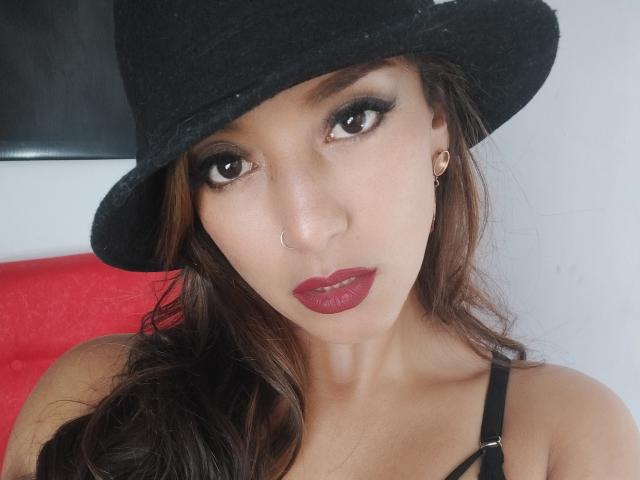 Hotcaroline33, con occhi marrone scuro, indossa un cappello nero, rossetto rosso e un piercing al naso. Ha capelli castani ondulati, un orecchino a cerchio dorato e una cintura nera..