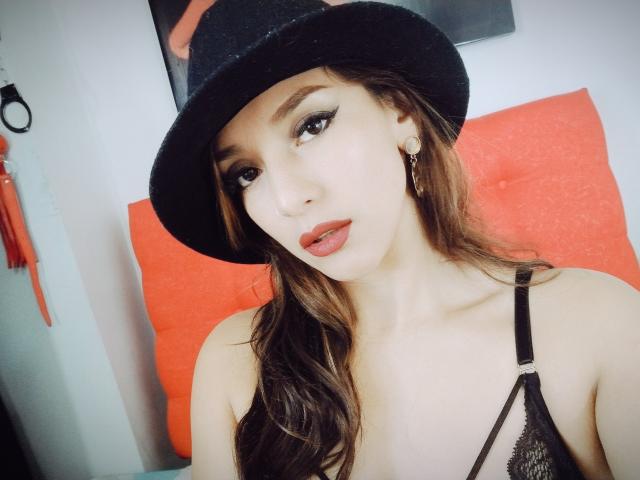 Hotcaroline33, avec de longs cheveux bruns, portant un chapeau noir, un soutien-gorge en dentelle noire et du rouge à lèvres, regarde l'appareil photo avec un léger froncement de sourcils, montrant ses seins..