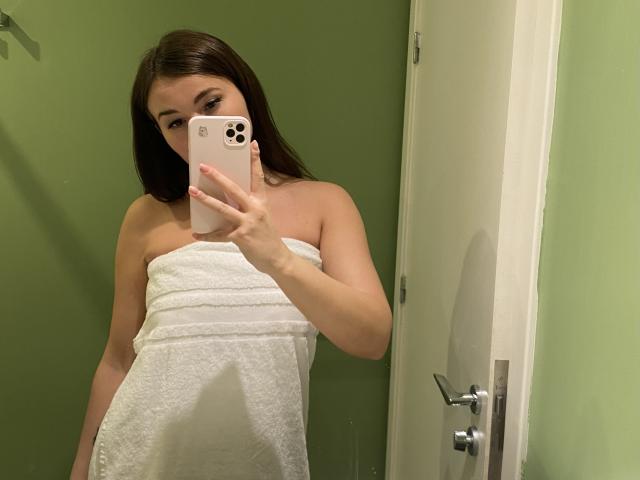 Dionisia with brown hair, wearing a white towel, holding a pink iPhone, covering her tits, taking a mirror selfie in a green-walled bathroom.