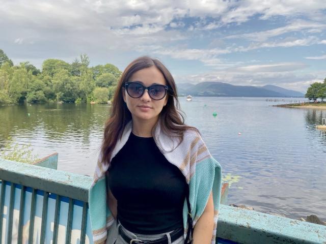 Dionisia with brown hair, wearing dark sunglasses, black top, and striped green-and-white scarf, standing by a blue railing, showing her ass with a black belt.