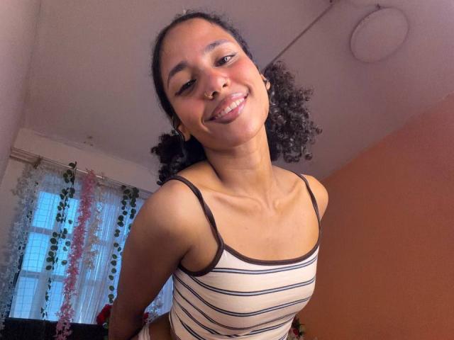 Cinthia Walker mit lockigem Haar, lächelnd, die Zunge herausstreckend, trägt ein gestreiftes Tanktop, zeigt ihre kleinen Brüste, hat eine Nasenring und Ohrringe und steht in einem sanft beleuchteten Raum..