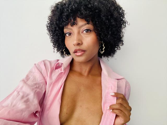 RoldanStefy with curly black hair, wearing a pink shirt, open to expose her tits, looking at the camera, holding the shirt, wearing gold earrings, light brown skin.