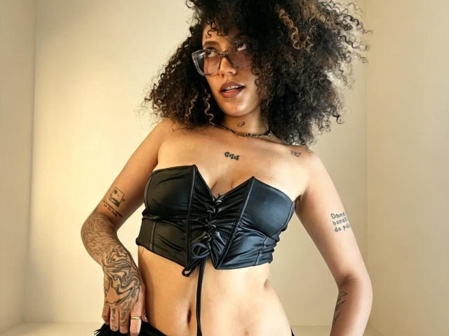 ScarlethVictoria with curly hair, wearing black strapless leather top, glasses, tattoos on arms, and a choker. Her tits are visible, and she's looking at the camera.