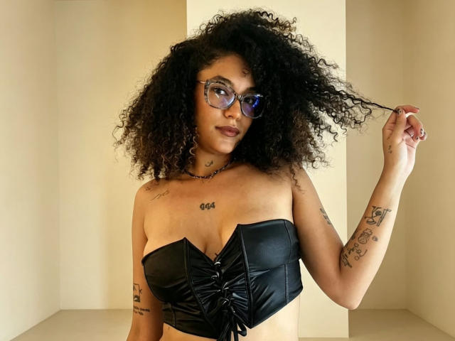 ScarlethVictoria with curly black hair, glasses, and black leather strapless top, holding her hair, showing tattoos, and a necklace, in a neutral-colored room. Boobs, tattoos, glasses.